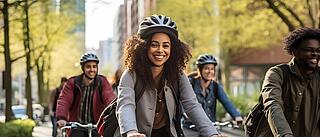 Eine Gruppe von Erwachsenen fährt gemeinsam mit Fahrrädern durch eine baumgesäumte Strasse in der Stadt. Im Vordergrund lächelt eine Frau mit lockigem Haar und Fahrradhelm in die Kamera. Die übrigen Radfahrer folgen ihr und wirken ebenfalls fröhlich.