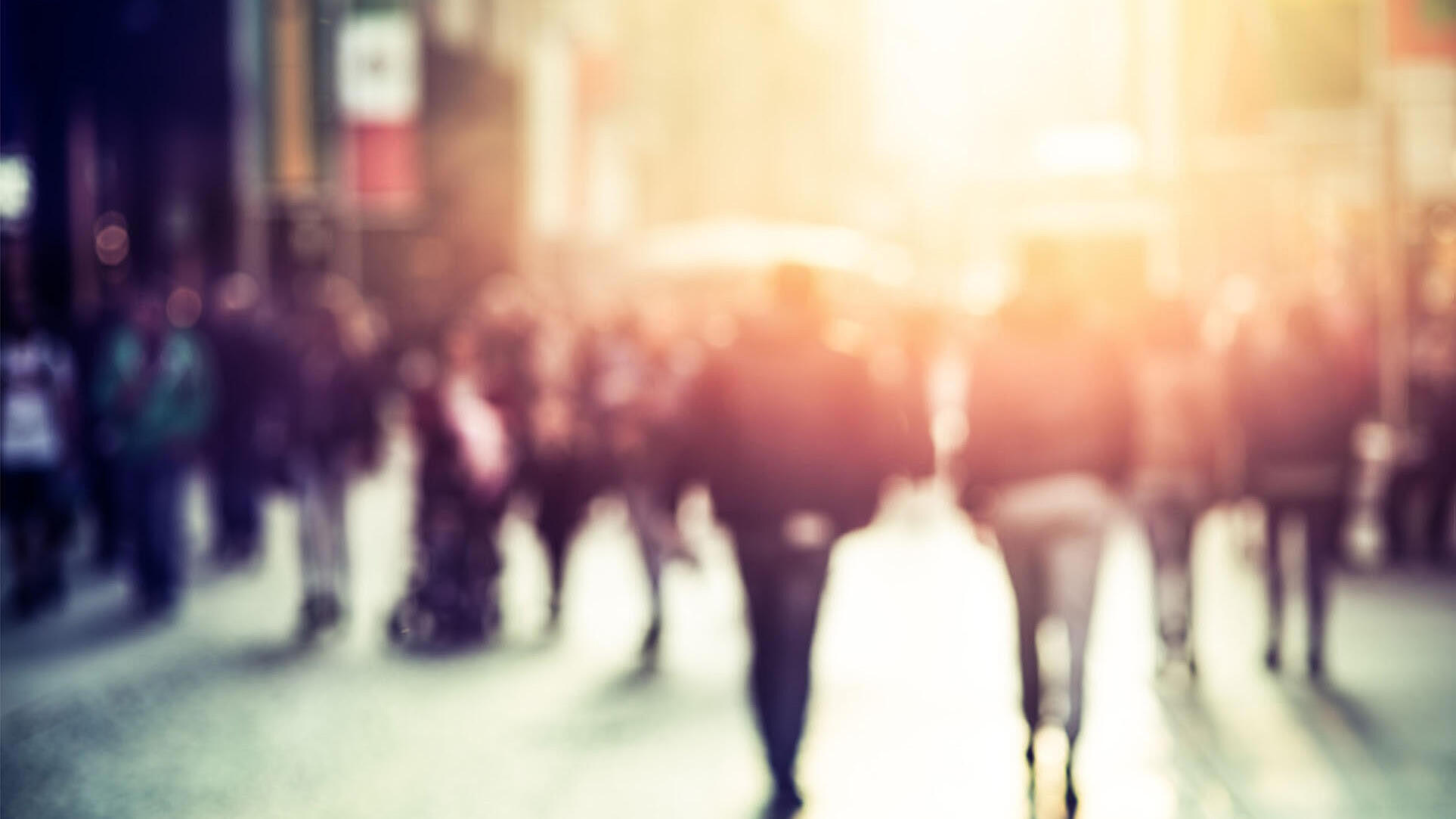 Unscharfes Foto einer Menschenmenge auf einer Stadtstraße. Der Hintergrund ist durch Sonnenlicht beleuchtet, die Personen sind nicht klar erkennbar.